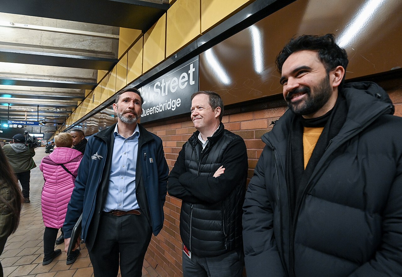Zohran Mamdani smiles alongside supporters in a New York subway station near a 63rd Street-Queensbridge campaign sign.