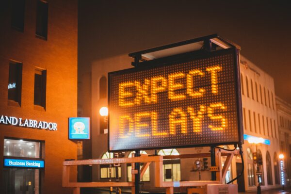 Electronic road sign at night displaying the message “Expect Delays” in bright orange lights on a city street.