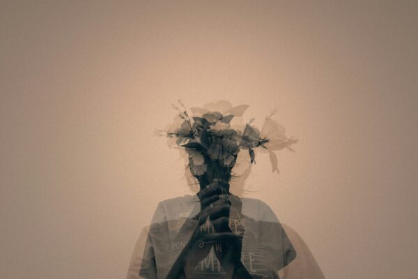 Double exposure image of a person holding a bouquet of flowers over their face, creating a ghostly, layered effect against a monochrome background, symbolizing the fragmentation of perception in schizophrenia.