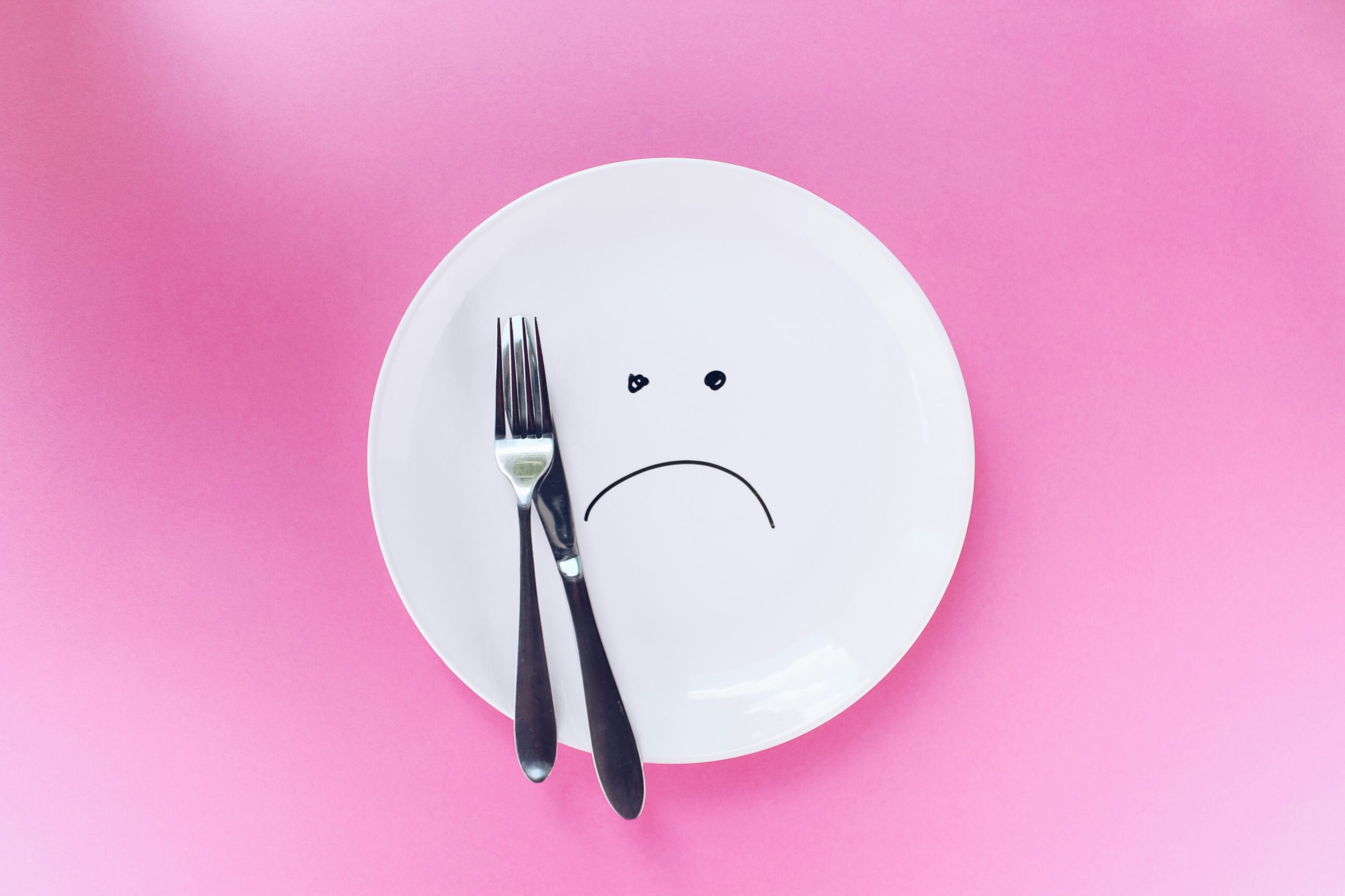 Empty white plate with a sad face drawn on it and utensils resting on top, symbolizing the emotional impact of hunger.