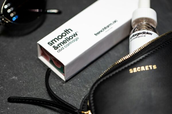 A CBD cartridge and sublingual oil placed beside sunglasses and a black leather purse on a dark countertop.