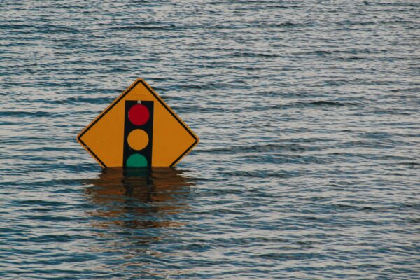 A partially submerged road sign with a traffic light symbol standing in floodwater, representing infrastructure failure and environmental risk