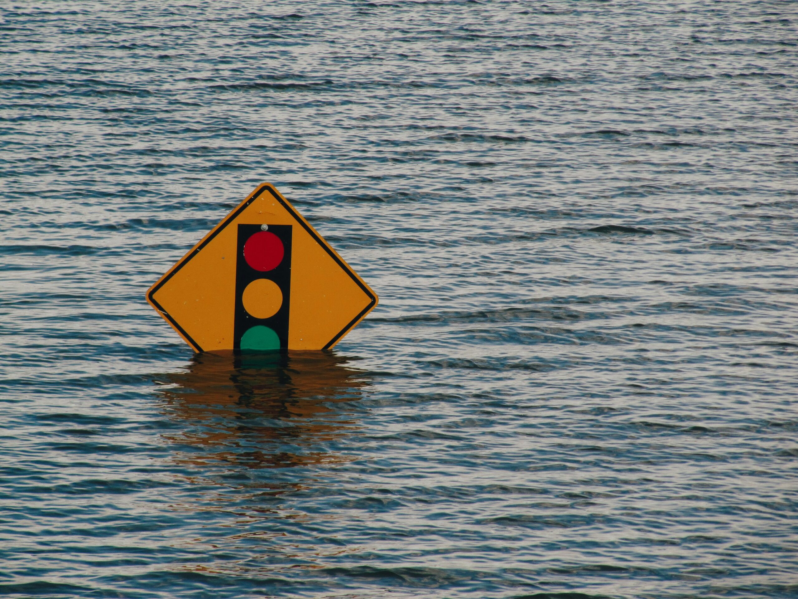 A partially submerged road sign with a traffic light symbol standing in floodwater, representing infrastructure failure and environmental risk