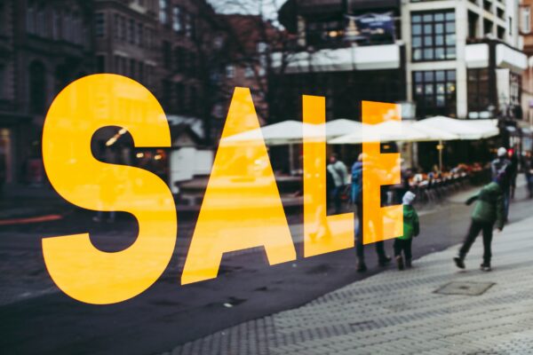 Large yellow ‘SALE’ sign on a store window during a busy shopping day, with shoppers walking by on a city street.