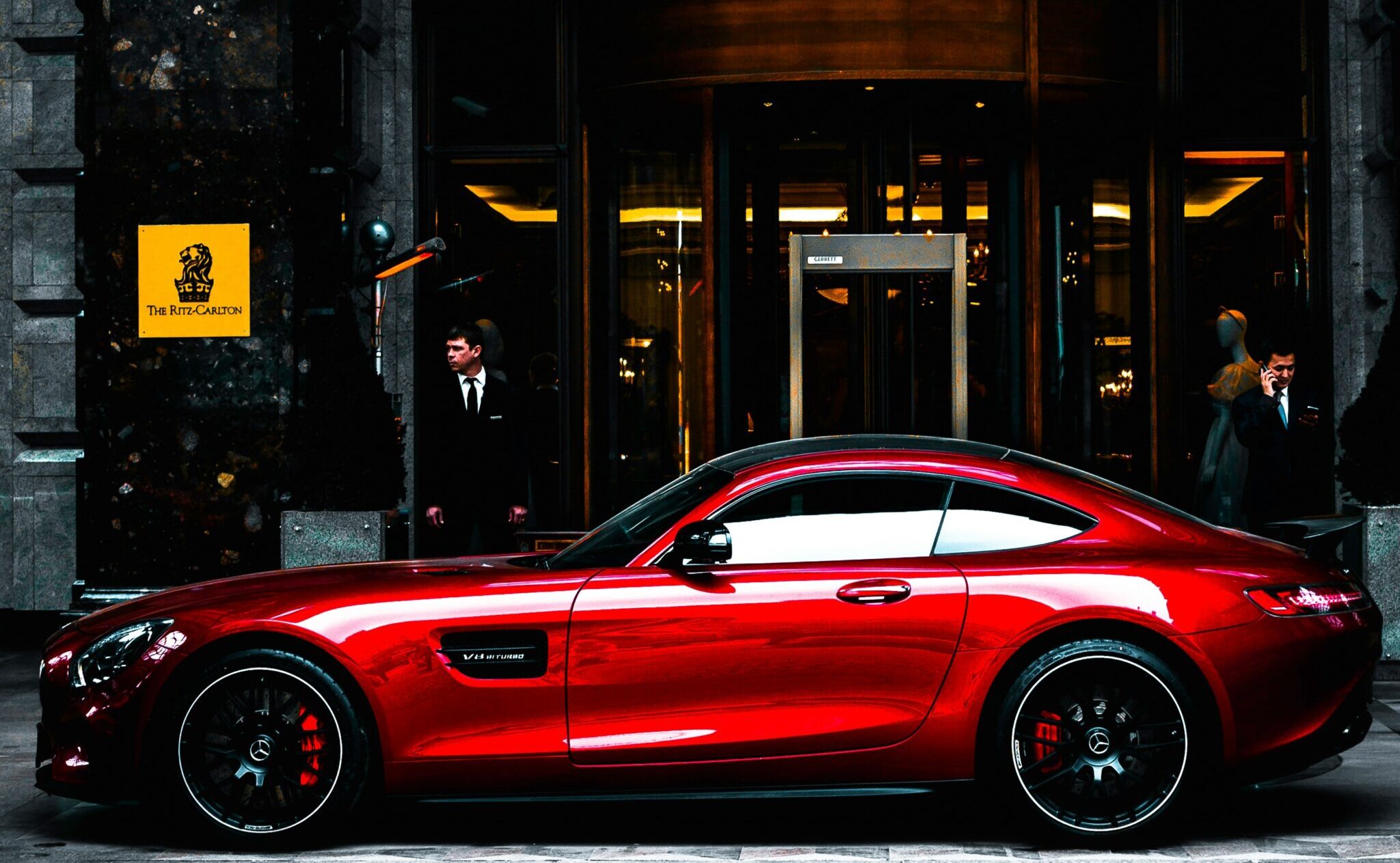 A bright red luxury sports car parked in front of an upscale hotel entrance with suited staff and elegant lighting in the background. All suggesting this man COULD be one of said billionaires.