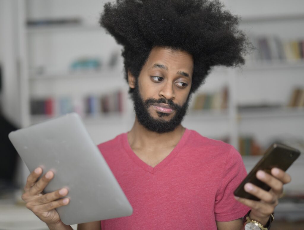 A man with afro hair multitasking with a tablet and smartphone indoors, showcasing technology use.