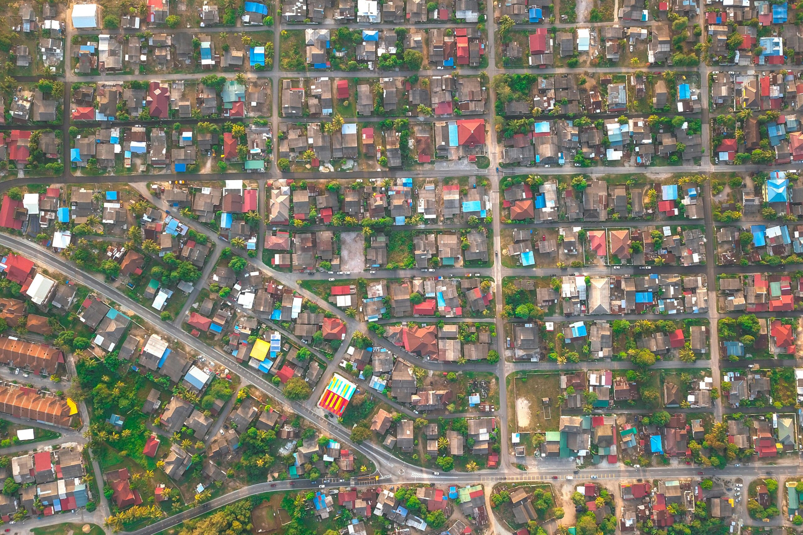 Top view of small multi colored cottages and housing surrounded by long narrow roads and vibrant green trees in daylight.