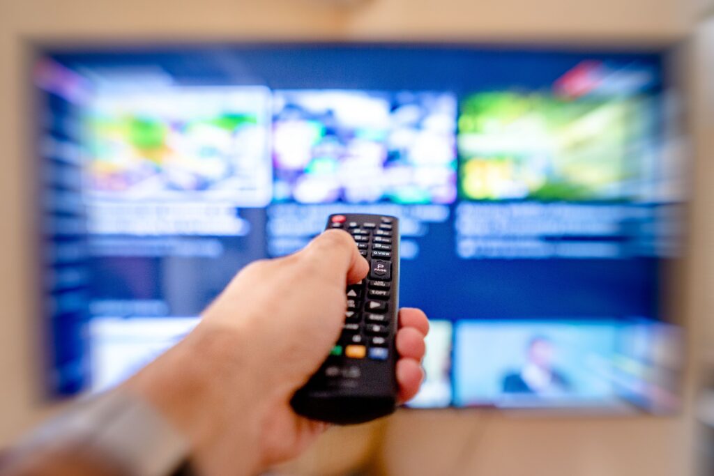 Hand holding remote control pointing at a blurred TV screen in a cozy indoor setting. Media Concentration.