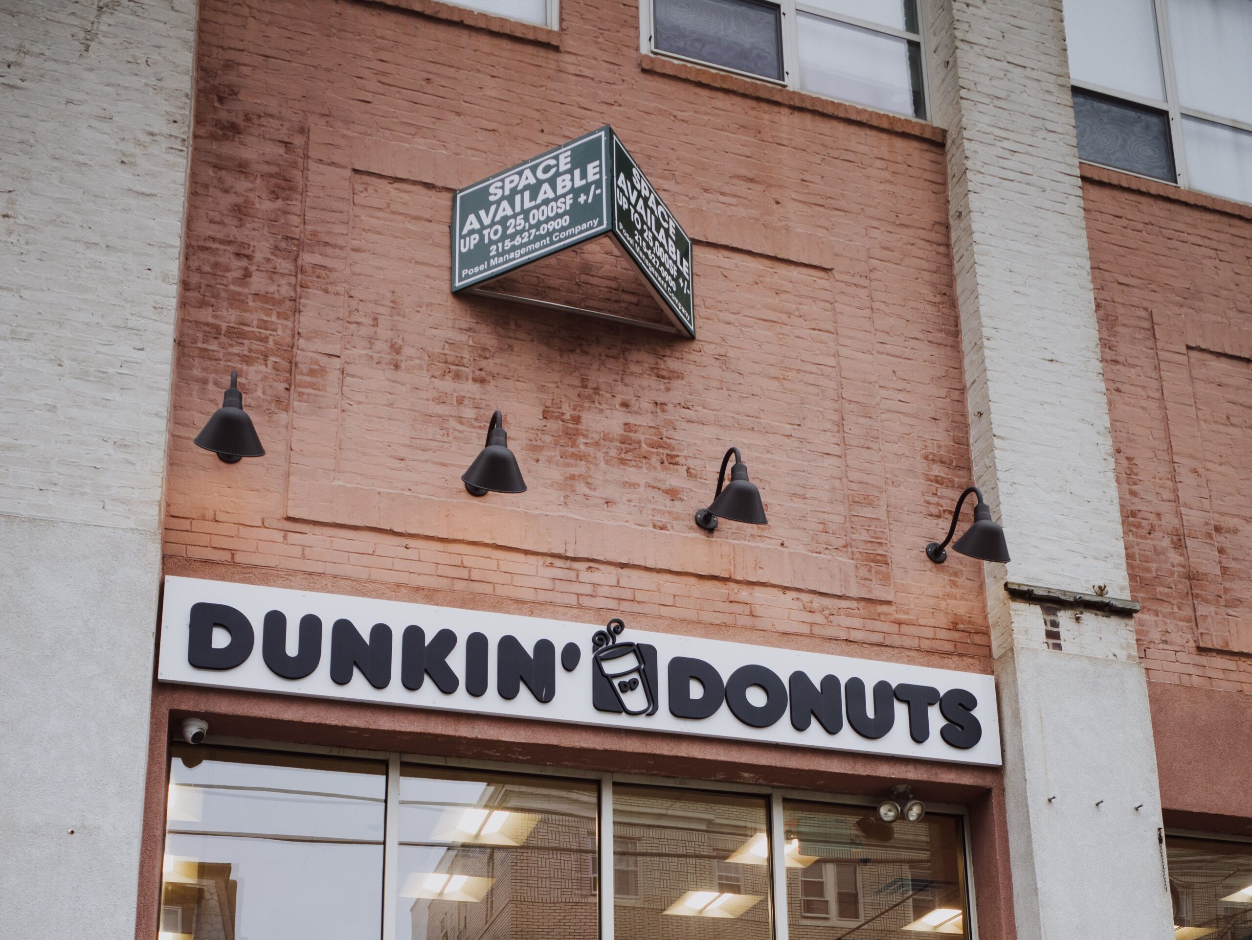 Man walking past Dunkin' Donuts, a fast food brand, with a space available sign on a city sidewalk, holding a drink.