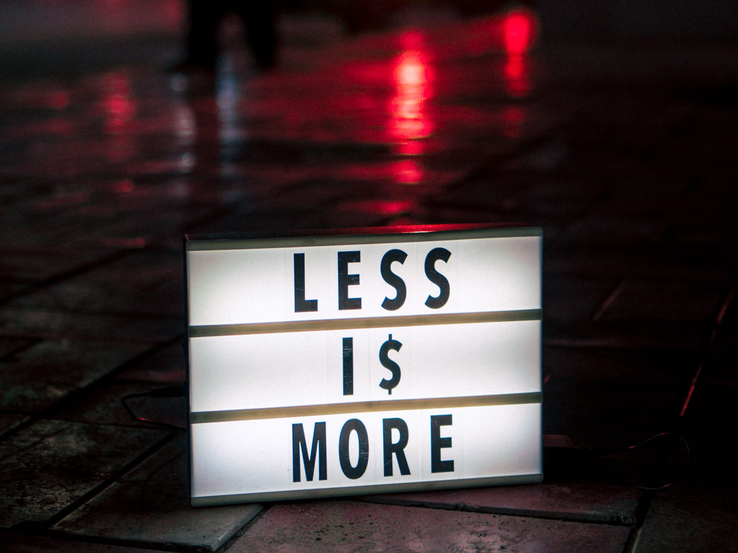 Minimalist lightbox sign reading 'Less is More' on a wet street at night, New Delhi.
