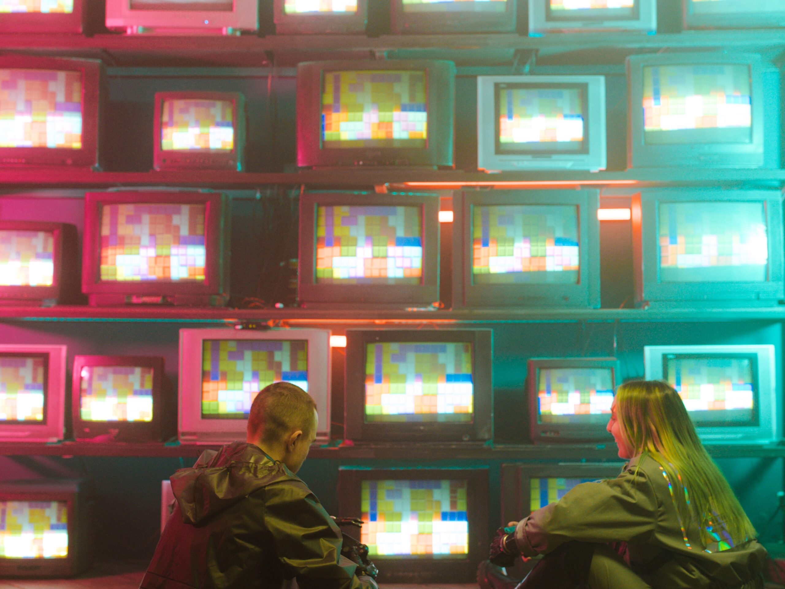Two gamers in futuristic attire sit by a neon-lit wall of retro TVs.
