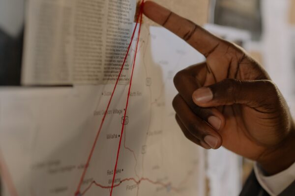A hand pointing at a map with red strings on a corkboard, depicting an investigation setup.