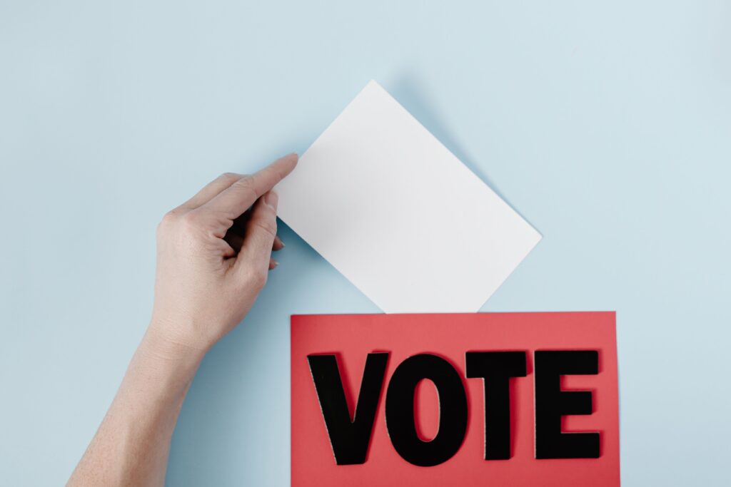 A hand places a ballot into a red box with the word VOTE against a blue background.