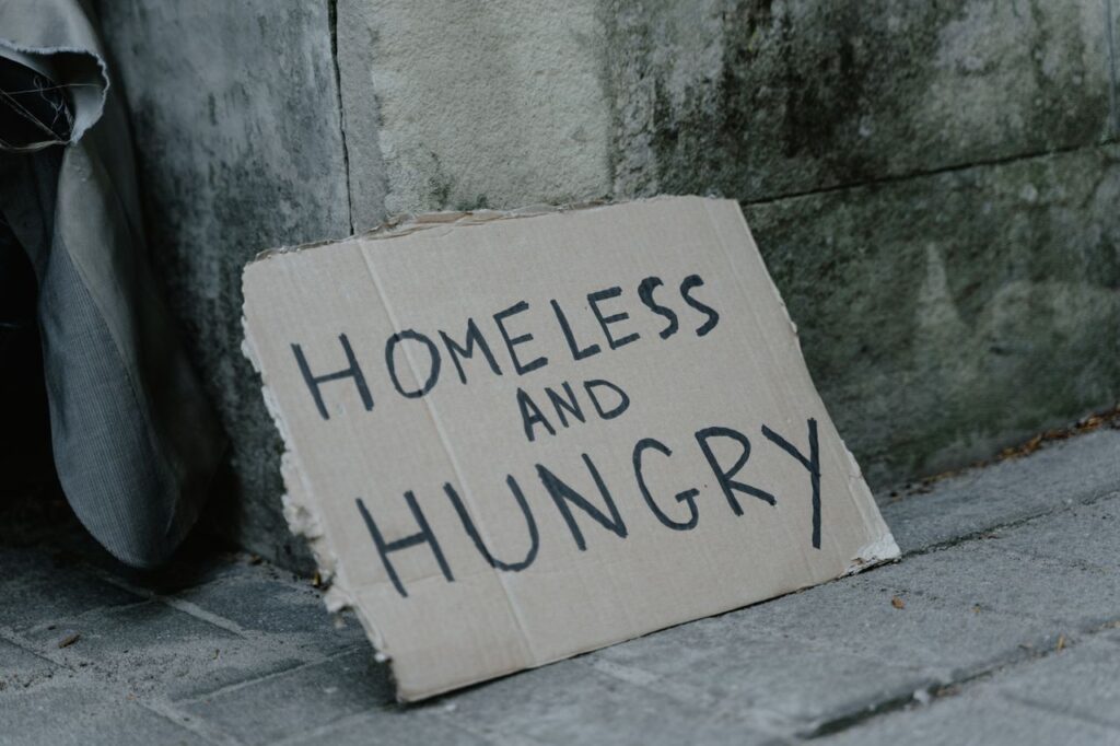 Cardboard sign with 'Homeless and Hungry' message leaning against a wall on a sidewalk.