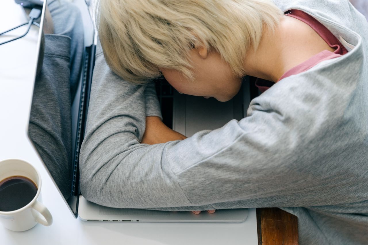 From above of anonymous female in casual clothes resting head on netbook keyboard near cup of coffee after long working day
