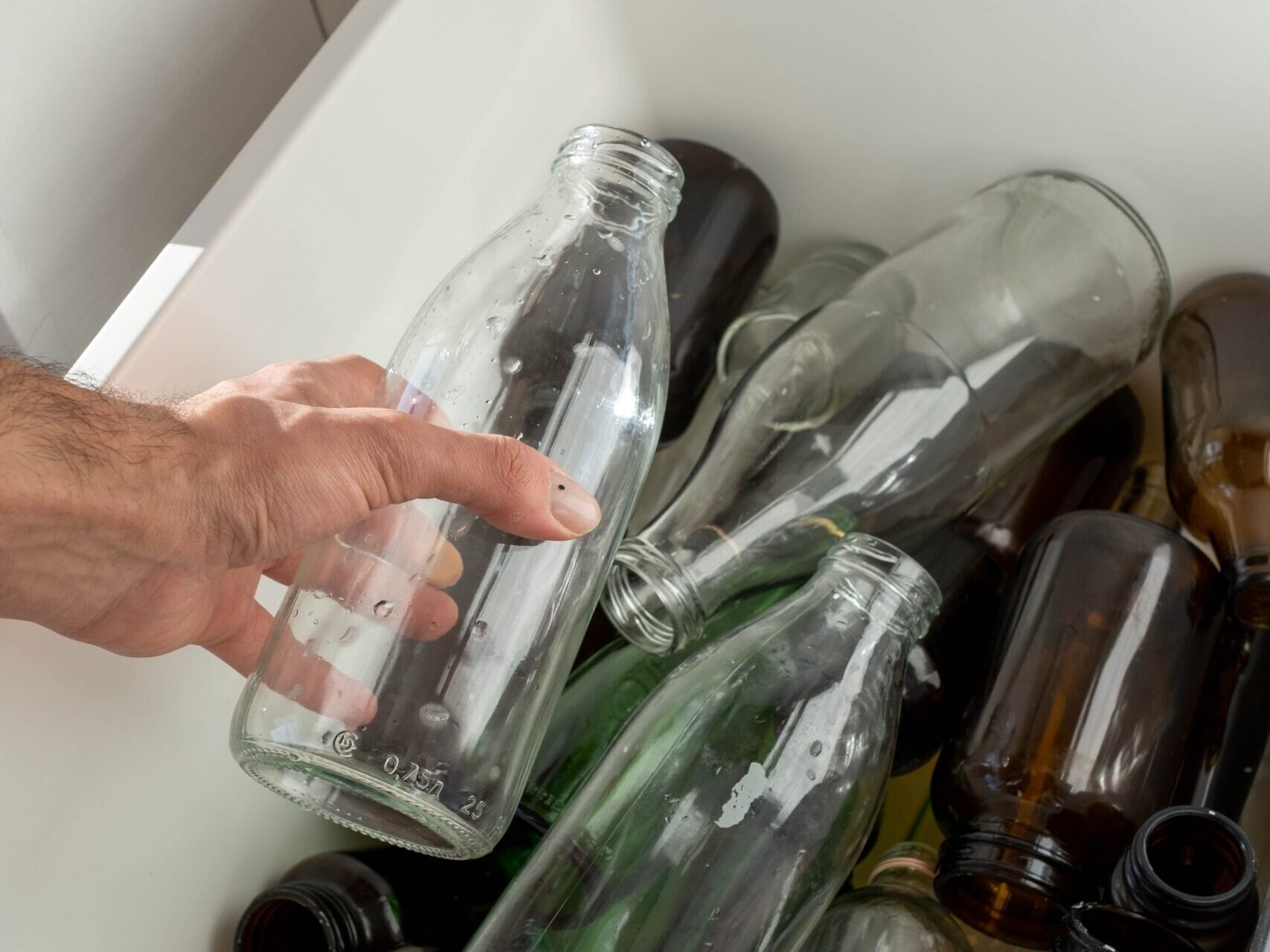A hand holds a glass bottle over a bin filled with various glass bottles for recycling.